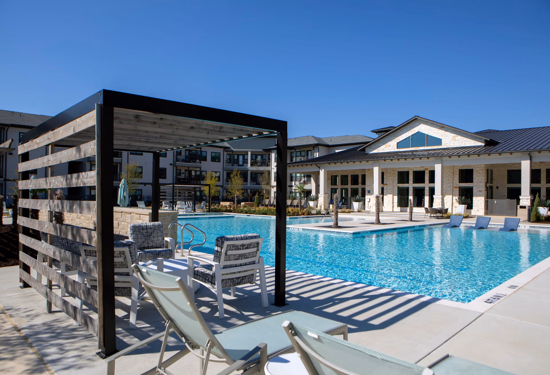 Outdoor swimming pool area at Solea Keller facility with lounge chairs and a shaded seating area under a pergola. The pool is surrounded by a concrete deck and modern buildings in the background under a clear blue sky.