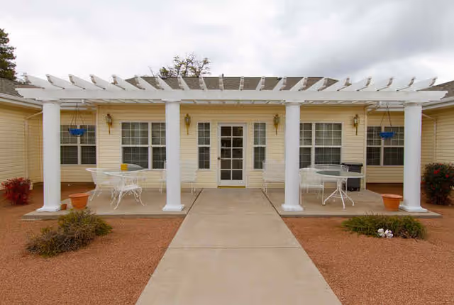Outdoor patio area at Avista Senior Living Payson featuring a white pergola with columns, two white metal tables with chairs, potted plants, and a concrete walkway leading to a glass door entrance of a yellow building with multiple windows.