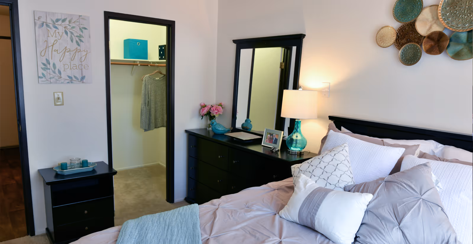 A neatly styled bedroom featuring a bed with decorative pillows, nightstands and lamp, a dresser with mirror, wall art, and an open walk-in closet.