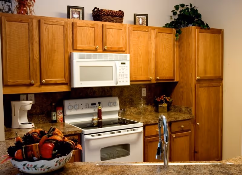 A kitchen with wooden cabinets, a white microwave mounted above a white electric stove, a coffee maker on the counter, a bowl of decorative vegetables on the counter, and a sink with a modern faucet in the foreground. There are decorative items on top of the cabinets including a basket, framed pictures, and a plant.