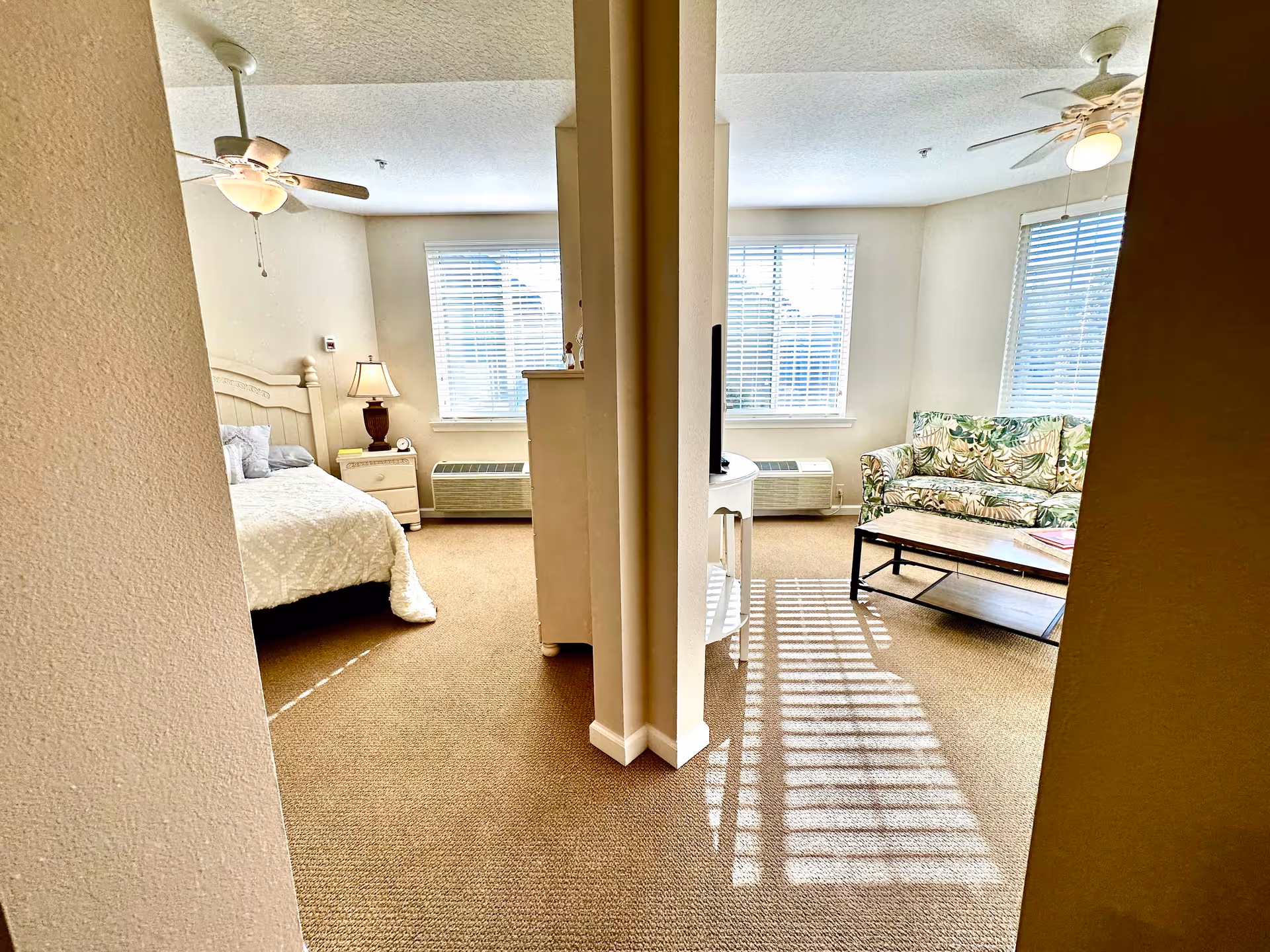 Interior view of a senior living facility unit showing a bedroom on the left with a bed, nightstand, lamp, and ceiling fan, and a living room on the right with a floral-patterned couch, coffee table, TV on a stand, ceiling fan, and windows with blinds letting in sunlight.