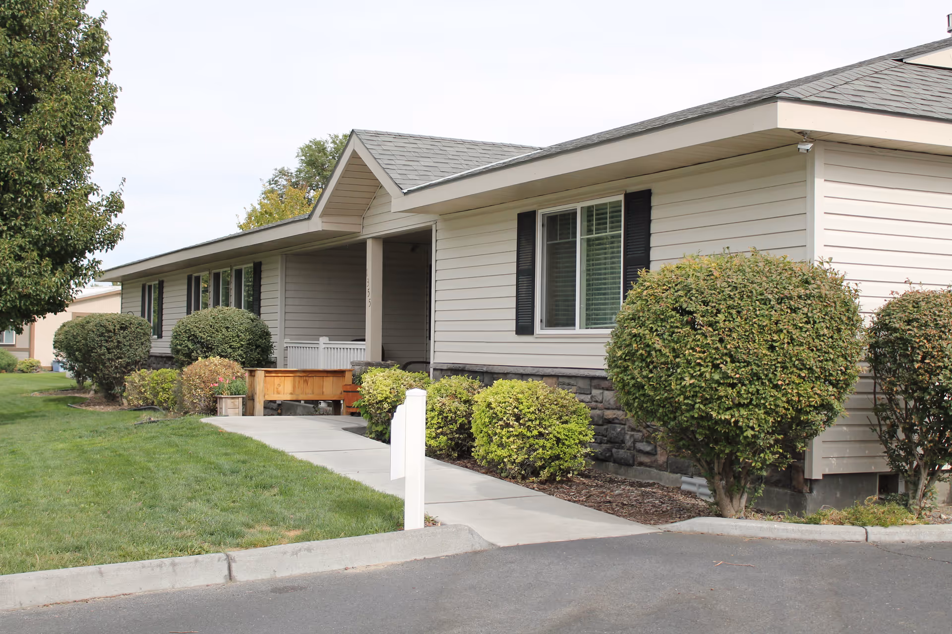 Exterior view of a single-story senior living facility building with beige siding, black shutters, and a gray shingled roof. The building is surrounded by neatly trimmed bushes and a well-maintained lawn, with a concrete walkway leading to the entrance.