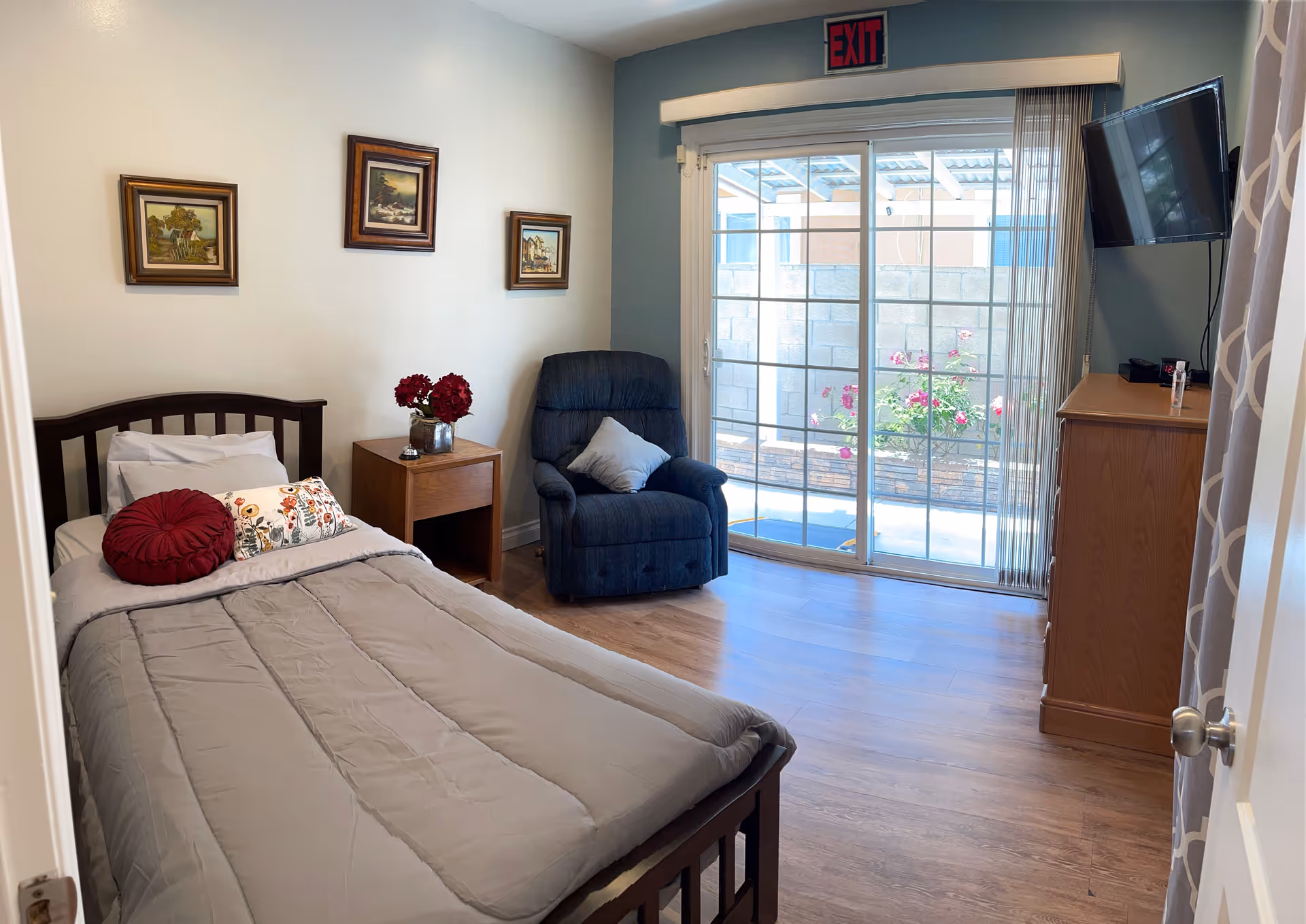 A cozy bedroom with a single bed covered in a gray comforter and decorative pillows. Next to the bed is a wooden nightstand with a vase of red flowers. A comfortable blue recliner chair with a gray pillow is positioned near a large sliding glass door that leads to an outdoor area with flowers. A wooden dresser with a mounted flat-screen TV above it is on the right side of the room. Three framed paintings hang on the wall above the bed. An exit sign is visible above the sliding door.