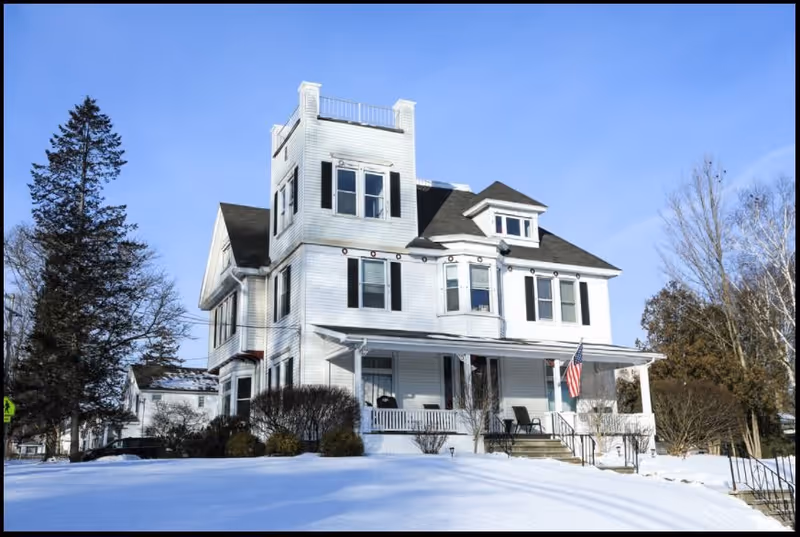 A large white multi-story house with black shutters and a wrap-around porch, surrounded by snow-covered ground and leafless trees under a clear blue sky. An American flag is displayed on the porch.