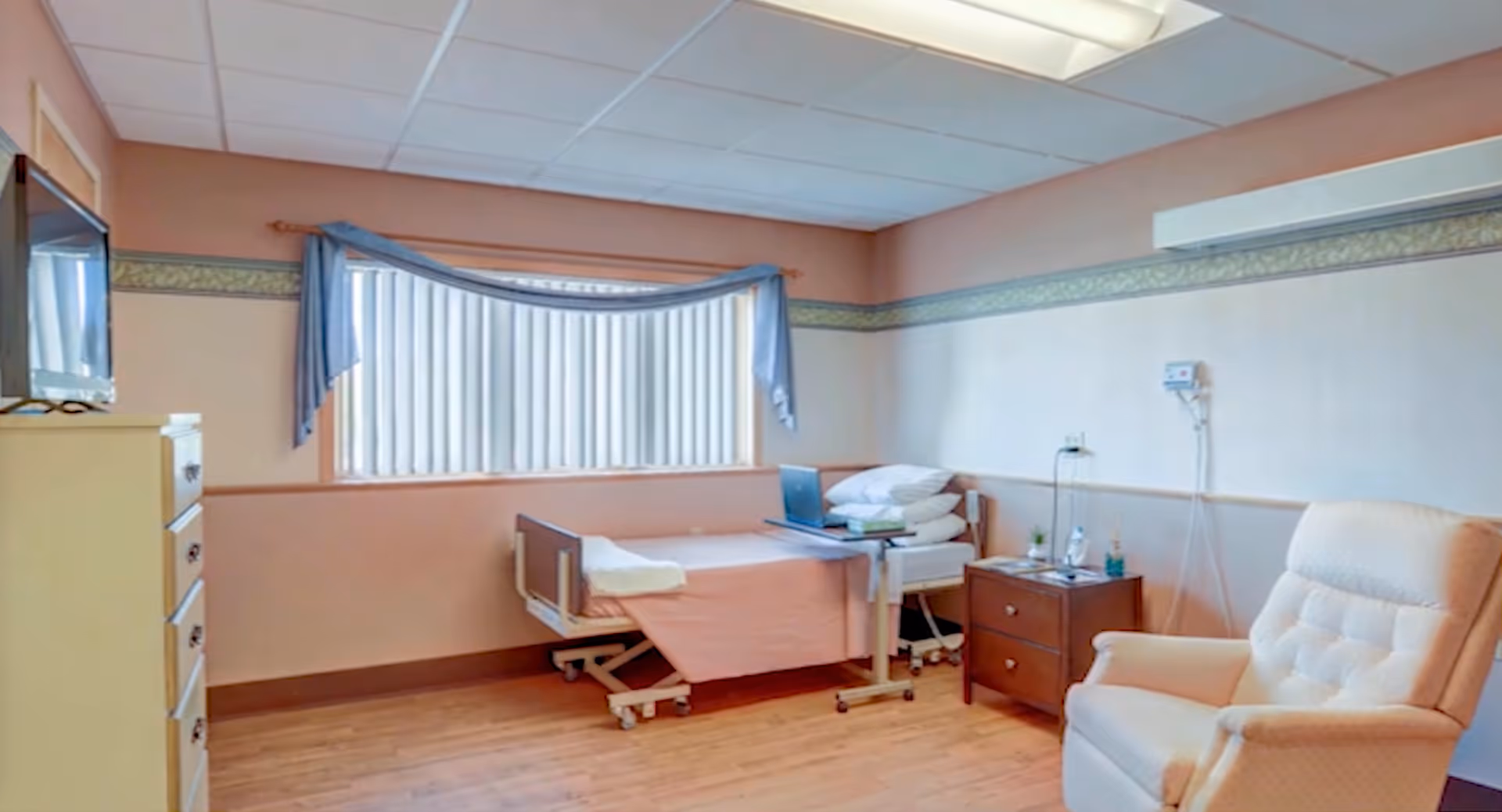 A healthcare facility room with a hospital bed, a small bedside table with items on it, a comfortable upholstered armchair, a chest of drawers with a TV on top, and a large window with vertical blinds and a blue valance.