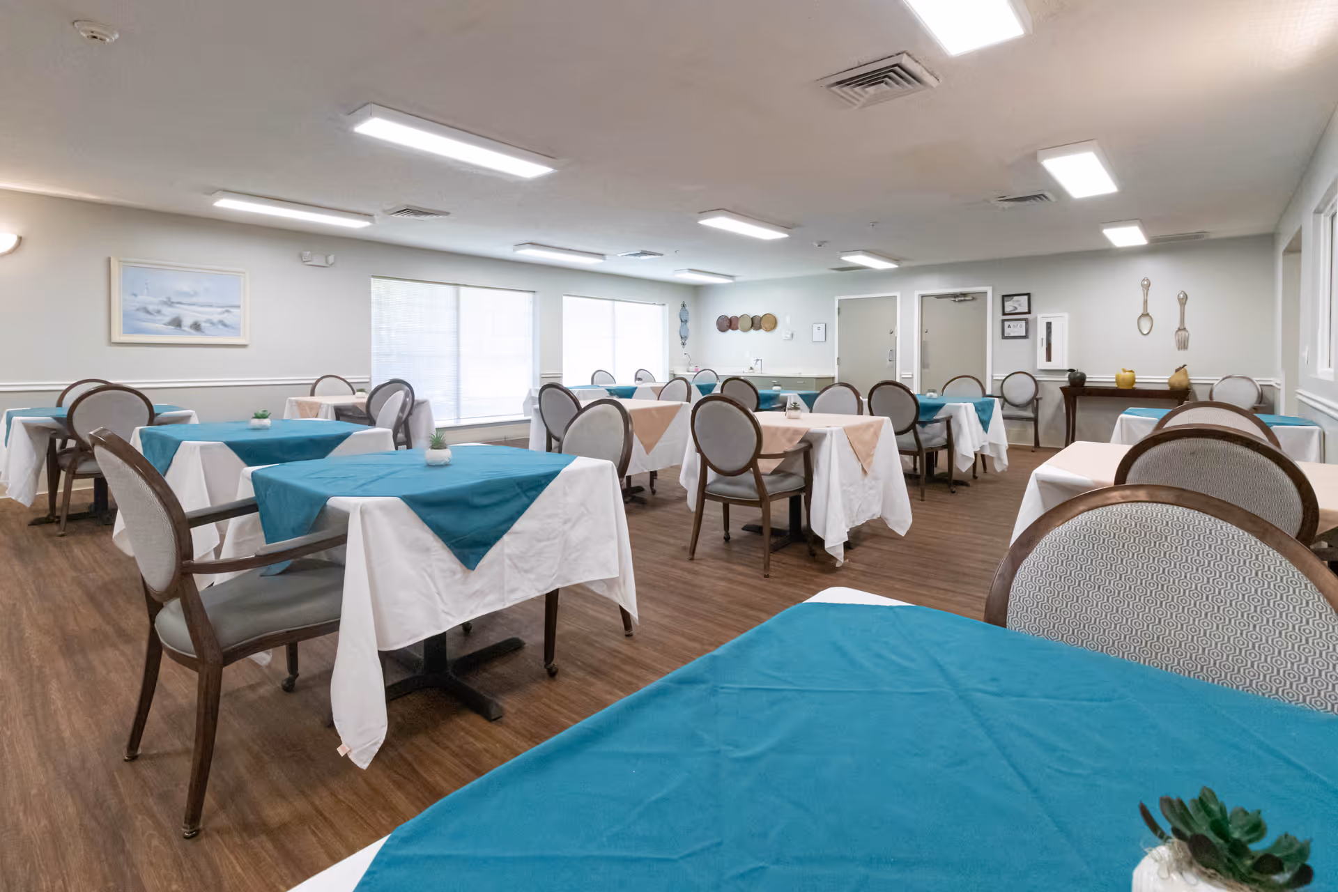 A dining room with multiple tables covered with white tablecloths and teal table runners. Each table is surrounded by cushioned chairs with wooden frames. The room has wood flooring, large windows with blinds, and neutral-colored walls decorated with framed artwork and wall hangings.