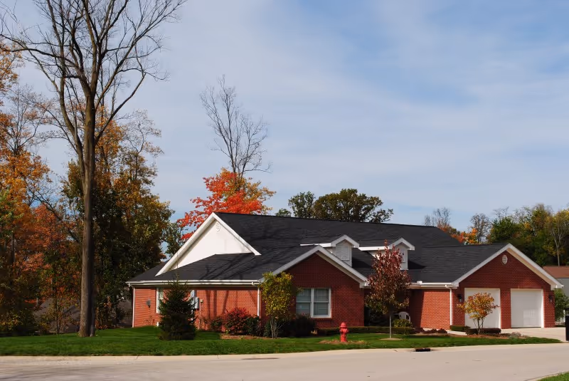 A single-story brick building with a black roof surrounded by trees with autumn foliage under a clear blue sky. The building has white trim and a garage on the right side. There is a fire hydrant and a well-maintained lawn in front.