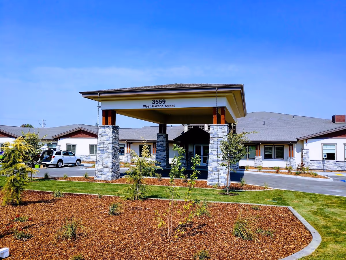 Front exterior view of a senior living facility with a covered entrance supported by stone pillars. The building is single-story with a gray roof and white walls accented with stone and wood. There are small trees and landscaped areas with mulch and grass in front of the building. A white SUV is parked near the entrance under a clear blue sky.
