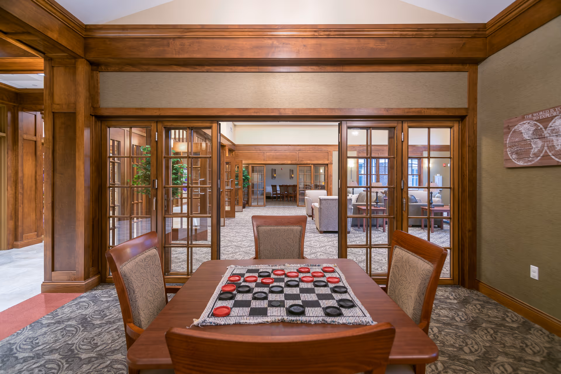 A cozy indoor seating area with a wooden table and four upholstered chairs, featuring a checkers game set up on the table. The room has wooden framed glass doors that open to a larger common area with sofas and additional seating. The walls are decorated with a world map artwork, and the space has warm lighting and carpeted floors.
