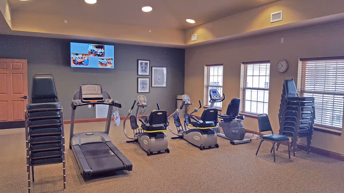 Exercise room with several pieces of fitness equipment including a treadmill and recumbent bikes. Two stacks of chairs are placed on either side of the room. There are windows with blinds on the right wall and a TV mounted on the far wall above framed pictures.