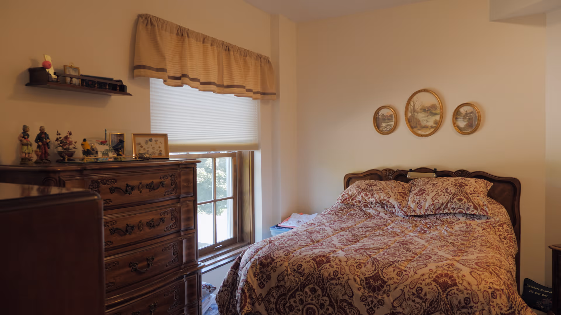 A cozy bedroom with a double bed covered in a patterned bedspread and matching pillows. Above the bed are three framed oval pictures. To the left of the bed is a wooden dresser with decorative items and framed photos on top. A window with a beige valance and a white blind is next to the dresser, letting in natural light.