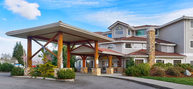 Exterior view of Creekside Continuing Care Community building with a covered entrance supported by wooden beams and stone pillars, surrounded by landscaped bushes and trees under a partly cloudy sky.