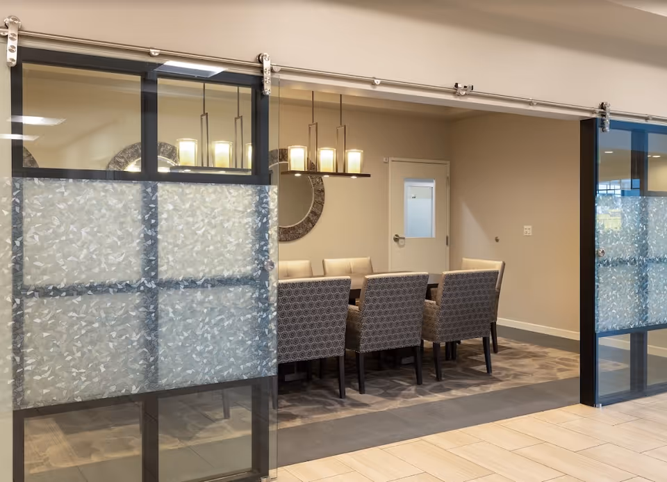 A modern dining room with a rectangular table surrounded by eight upholstered chairs. The room is enclosed by sliding glass doors with frosted patterns on the lower half. A contemporary light fixture with multiple lights hangs above the table, and a round decorative mirror is mounted on the beige wall in the background.