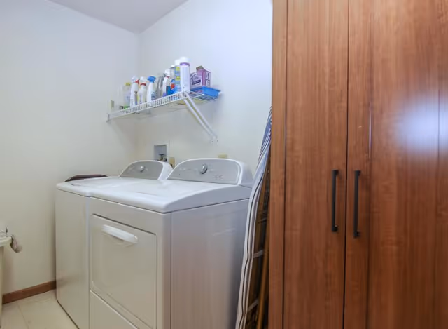 Laundry room with a white washing machine and dryer side by side, a wooden cabinet on the right, an ironing board leaning against the wall, and a shelf above holding various cleaning supplies and laundry products.