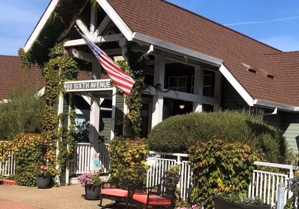 Exterior view of a building with a peaked roof covered in brown shingles. The entrance is framed by white wooden beams with green ivy climbing on them. An American flag is hanging near the entrance. There are potted plants with flowers and two black chairs with red cushions in front of the building. A sign above the entrance reads '900 SIXTH AVENUE'.