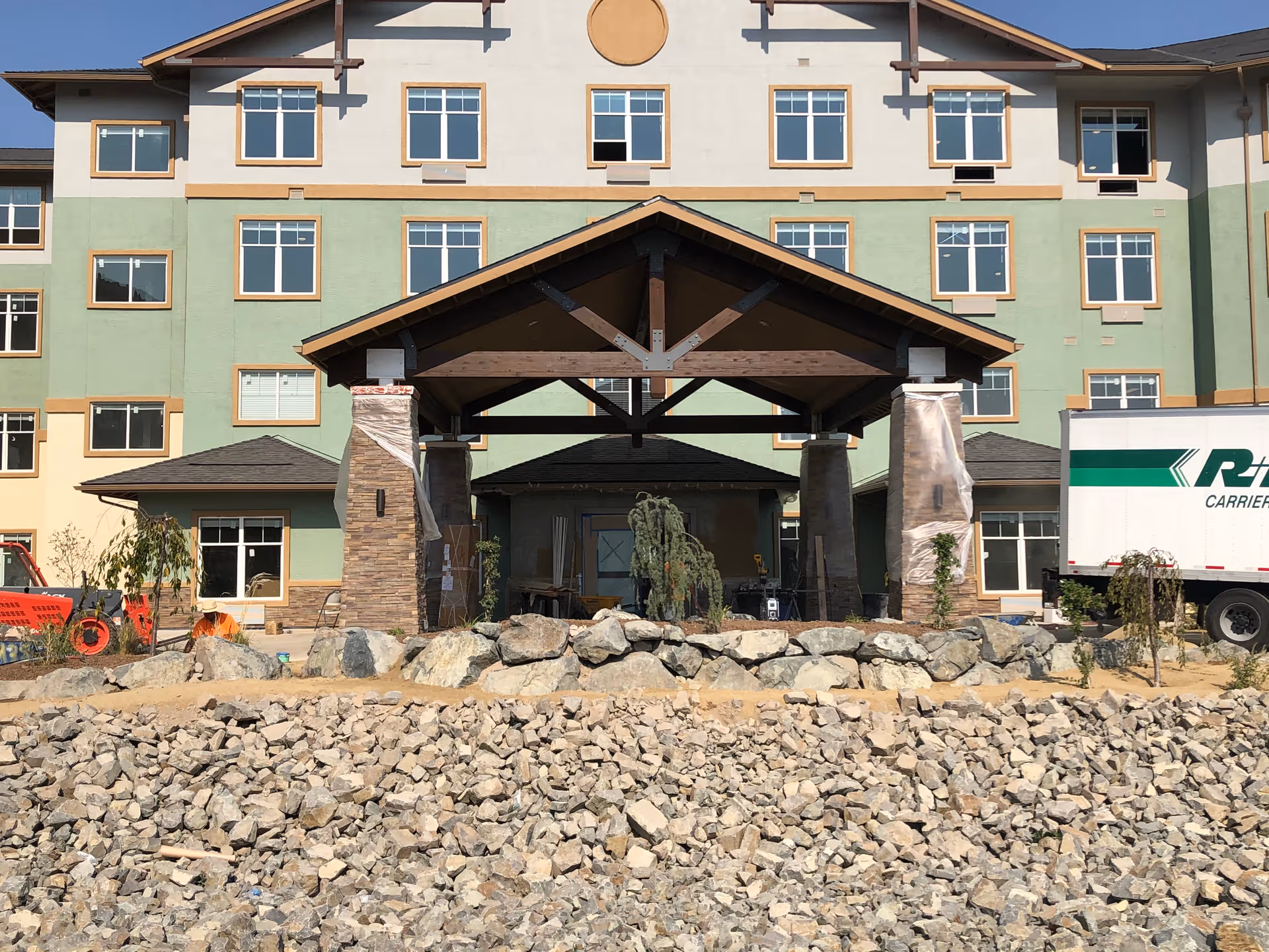 Front exterior view of a multi-story building under construction with a covered entrance supported by stone pillars. There are rocks and construction materials in the foreground, and a truck is parked on the right side.