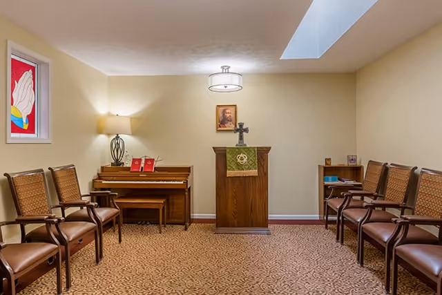 Small chapel-style room with rows of chairs, a wooden podium topped with a cross, a piano, and a stained-glass window.