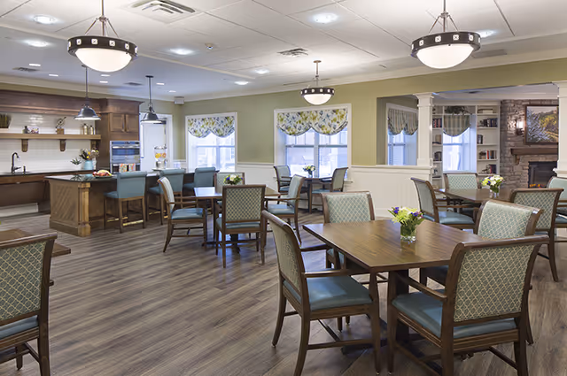 A bright and spacious dining area in a senior living facility with multiple wooden tables and cushioned chairs arranged neatly. The room features large windows with floral valances, pendant and ceiling lights, and a kitchen area with a counter and stools in the background. There is also a cozy seating area with a stone fireplace and built-in shelves.