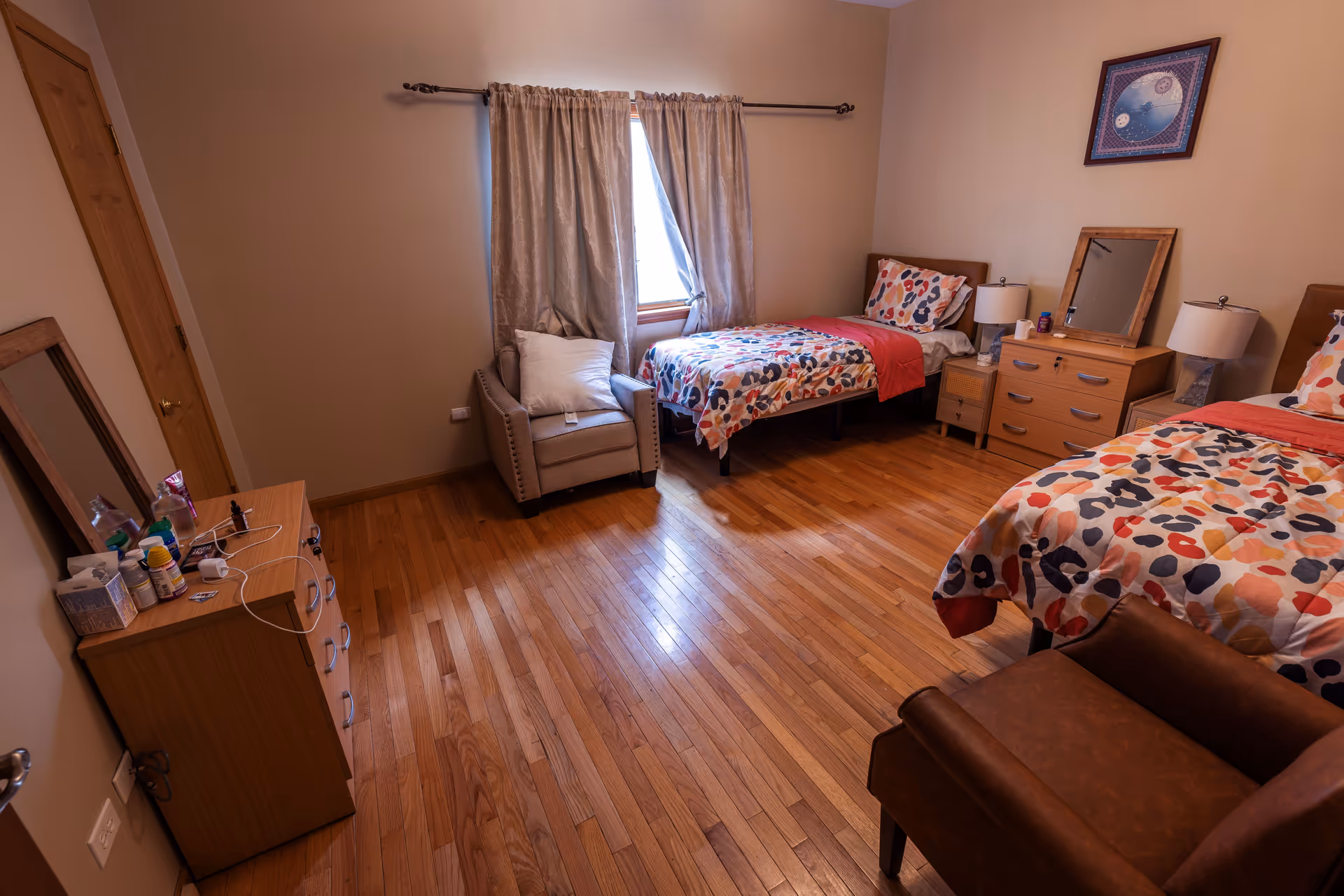 A senior living bedroom with two single beds covered in colorful patterned bedding. Between the beds is a wooden dresser with a mirror and two lamps. There is a window with beige curtains, a beige armchair with a pillow, and a wooden floor. Another dresser with personal items and a mirror is visible on the left side of the room.
