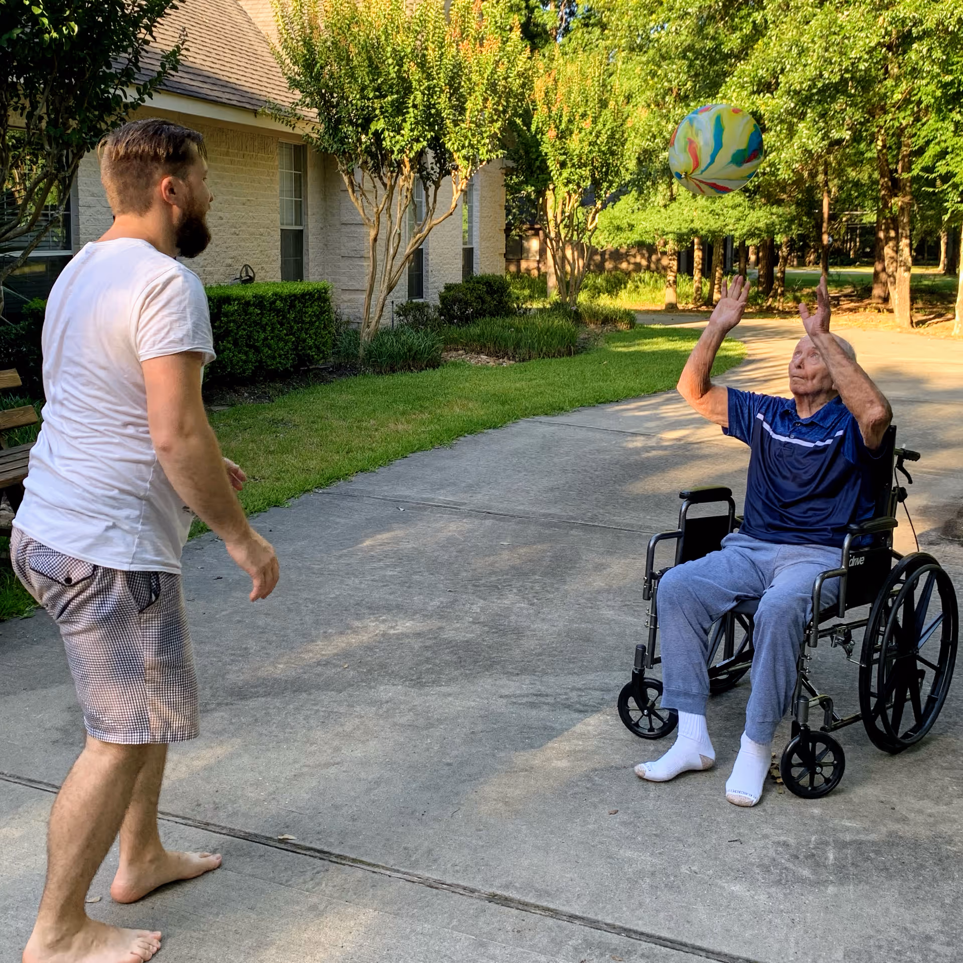 An elderly man in a wheelchair playing with a colorful beach ball outdoors on a paved pathway, accompanied by a younger man standing barefoot nearby. The scene is set in a green, tree-lined area next to a building.
