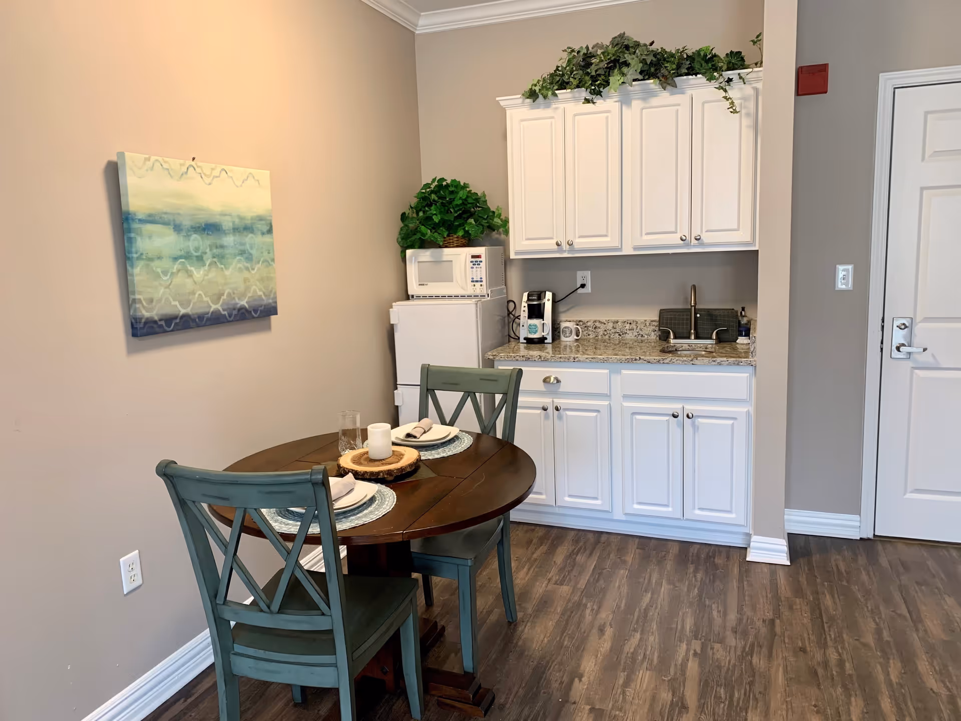 A small dining area with a round wooden table set for two with plates, napkins, and a candle centerpiece. Behind the table is a kitchenette with white cabinets, a granite countertop, a small sink, a coffee maker, a microwave, and a mini refrigerator. There is a green plant on top of the microwave and decorative greenery above the cabinets. The walls are painted beige, and there is a blue and green abstract painting hanging on the wall to the left.