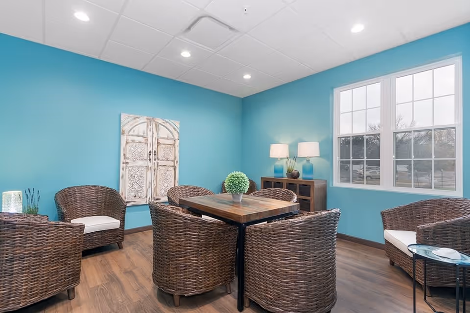 A cozy seating area with six wicker chairs arranged around a wooden table in a room with turquoise walls, a large window, wooden flooring, and a sideboard with two lamps and decorative plants.