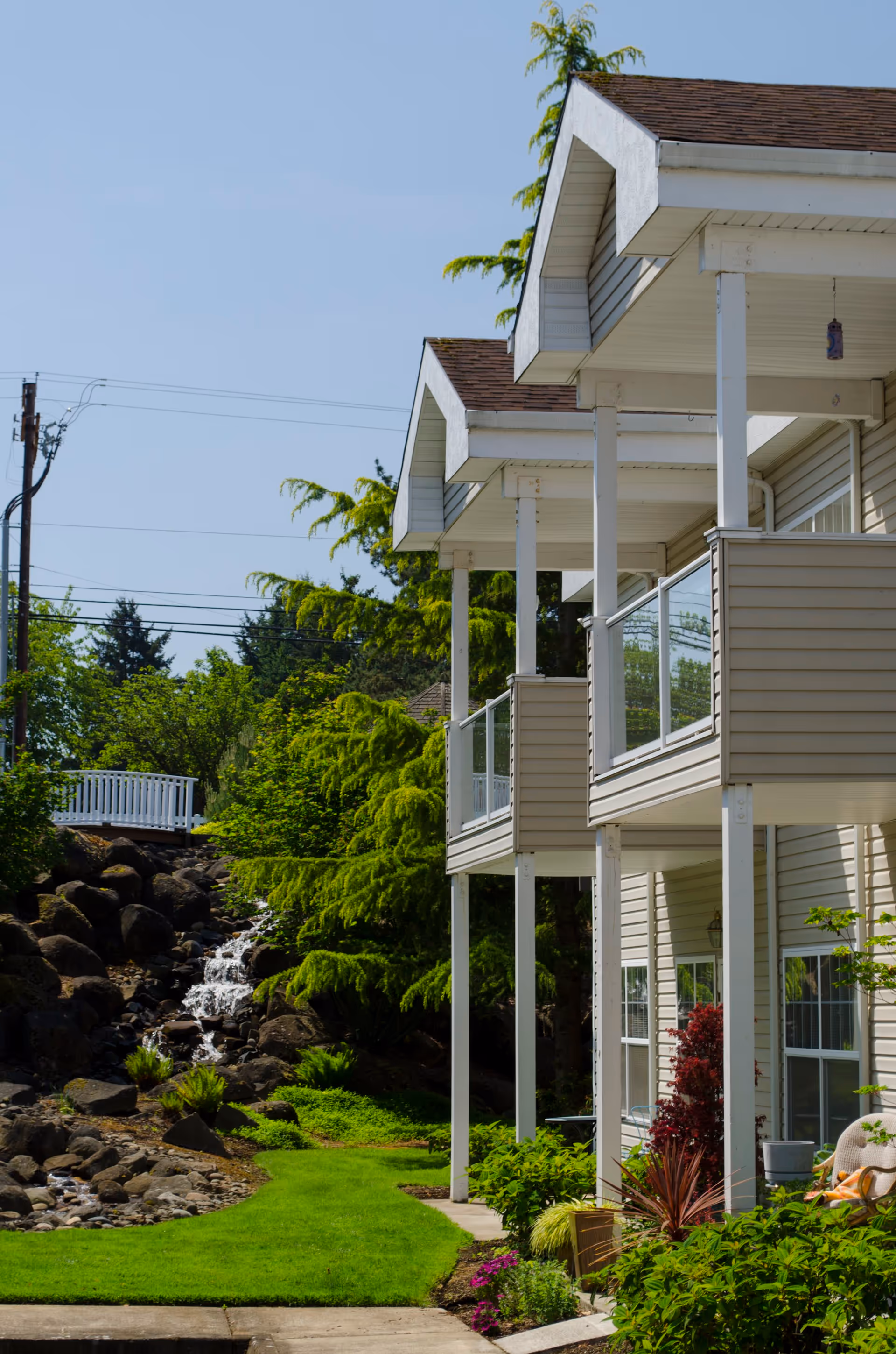 Side view of a residential building with beige siding and white trim, featuring balconies with glass railings. The building is surrounded by green shrubs, plants, and a small landscaped area with rocks and a small waterfall flowing down the rocks. The sky is clear and blue.