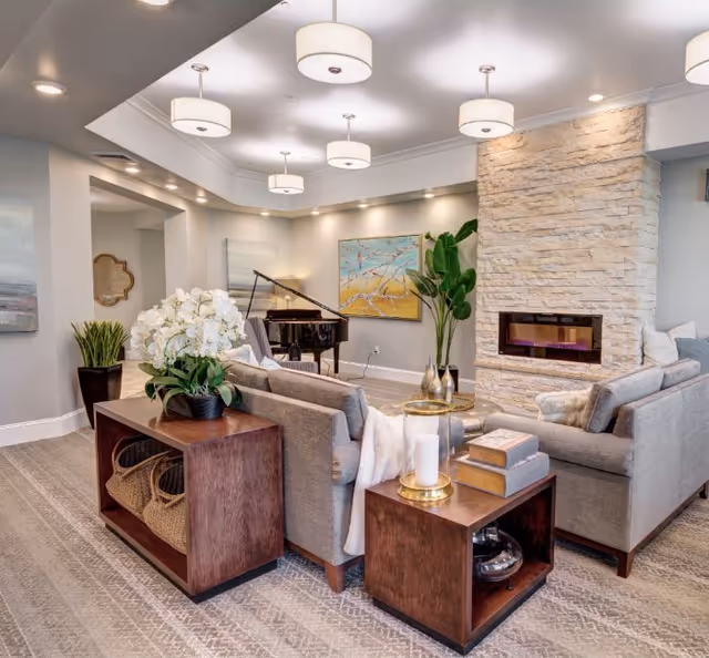 Contemporary communal living room with sofas arranged around a stone fireplace, a grand piano, pendant lights, plants, and decorative side tables.