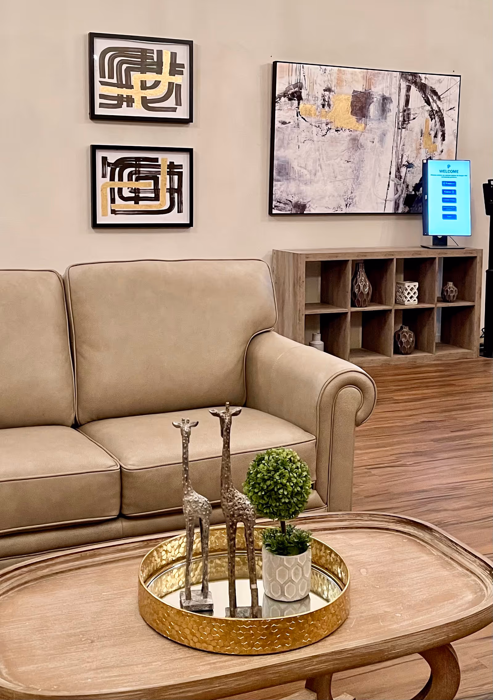 A beige leather sofa and wooden coffee table with decorative giraffe figurines and a small potted plant in a lobby-style living area with wall art and shelving behind.