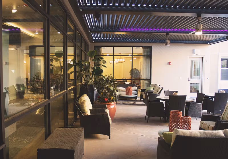 Covered outdoor patio area with wicker chairs, tables, and potted plants. The patio has a slatted roof with purple accent lighting. Large windows show an indoor seating area with sofas and a chandelier inside the building.