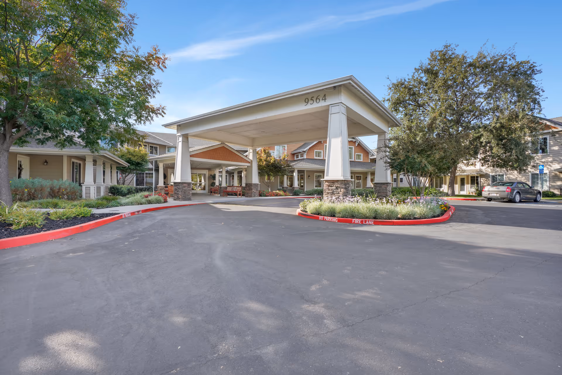 Entrance area of a senior living facility with a large covered drop-off area supported by four columns. The building has a beige exterior with stone accents and is surrounded by trees and landscaping. The address number 9564 is visible on the covered structure. A car is parked on the right side near the building.