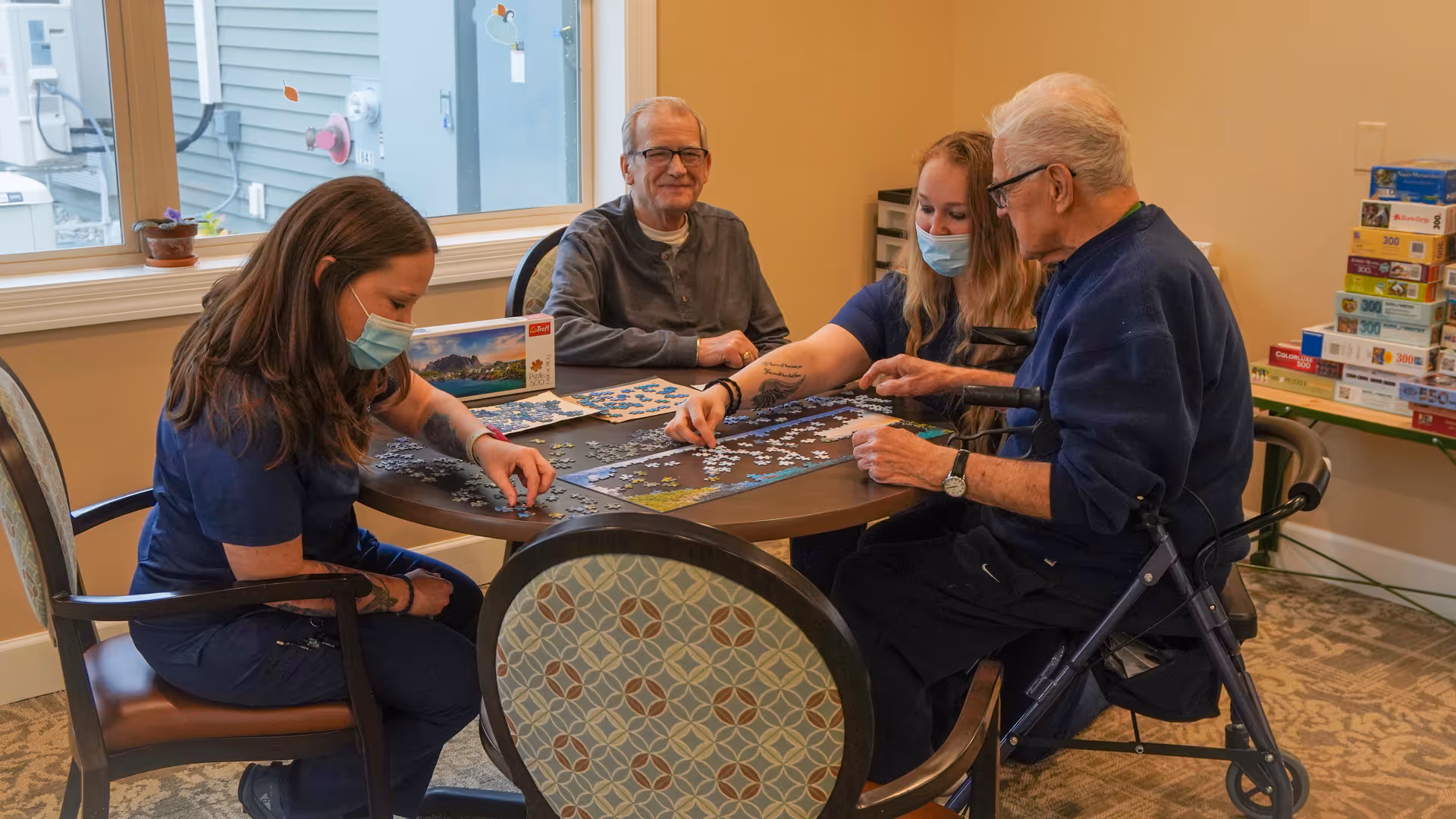Two elderly men and two women, one wearing a face mask, sitting around a round table working on a jigsaw puzzle in a well-lit room with a window and a stack of puzzle boxes in the background.