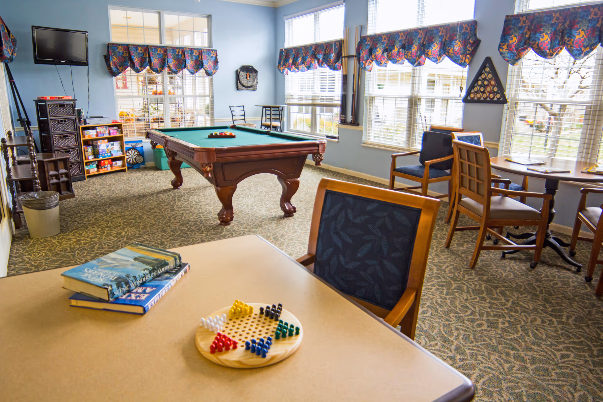 Bright senior recreation room with a pool table, tables and chairs, books and board games by large windows.