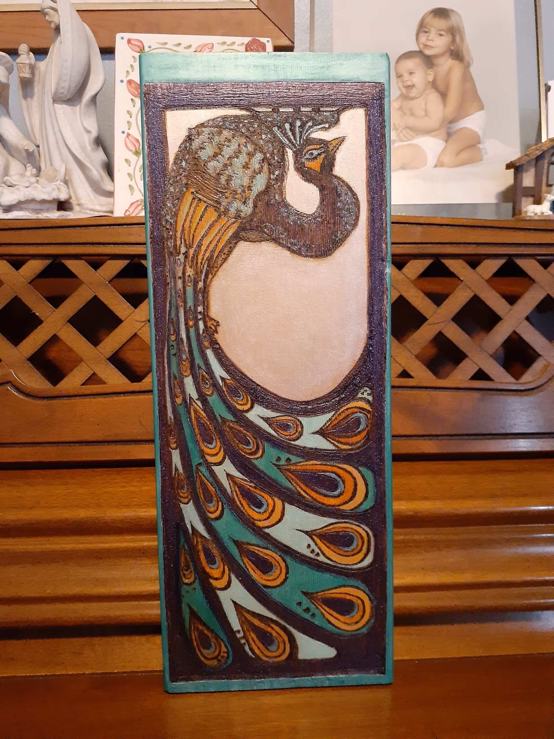 A painted wooden panel depicting a stylized peacock displayed on a wooden piano with figurines and a framed photo behind it.