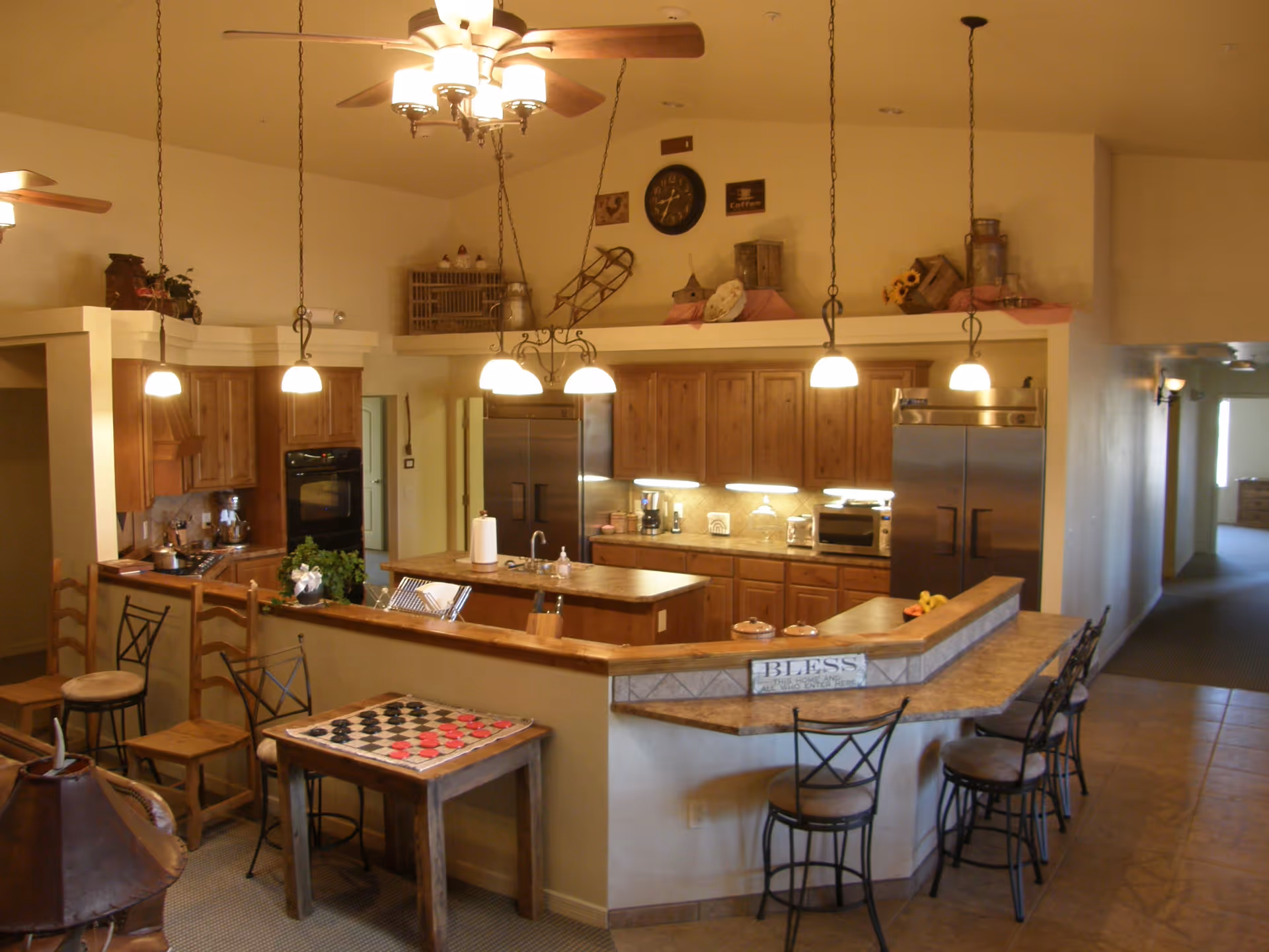 Interior view of a kitchen area with wooden cabinets, stainless steel appliances, and a countertop with bar stools. There are hanging pendant lights and ceiling fans with lights. A small table with a checkers game is visible in the foreground. Decorative items and a clock are placed on a shelf above the cabinets.