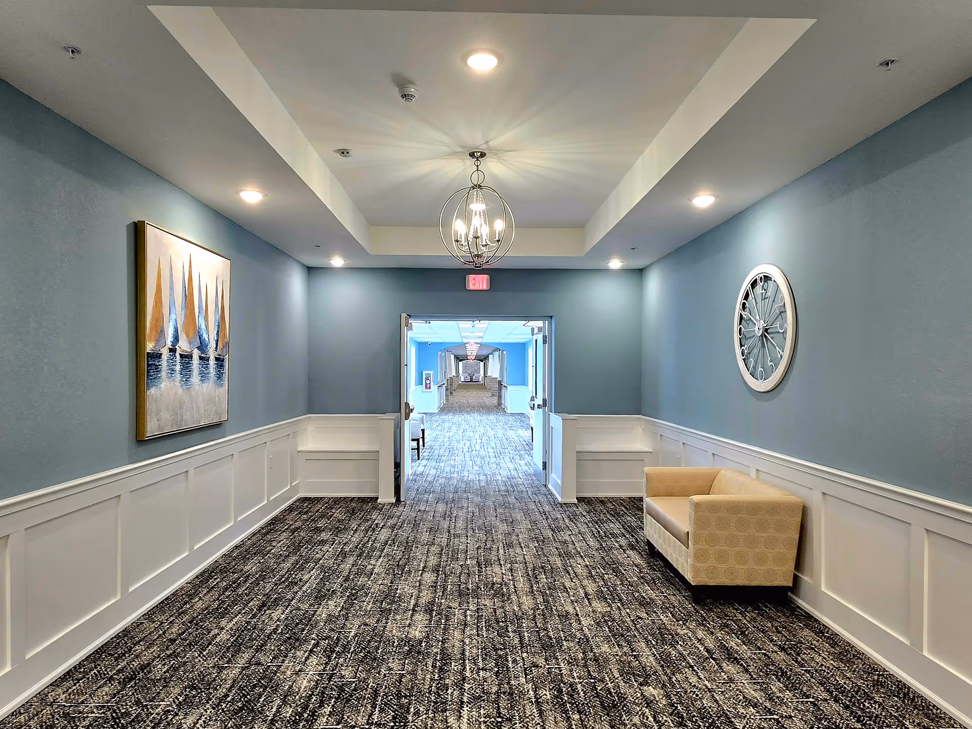 A well-lit hallway with blue walls and white wainscoting, featuring a beige patterned loveseat on the right side and a large round wall clock above it. On the left wall, there is a painting of sailboats. The hallway has a patterned carpet and a modern chandelier hanging from the ceiling. Double doors at the end open to another corridor with similar decor.
