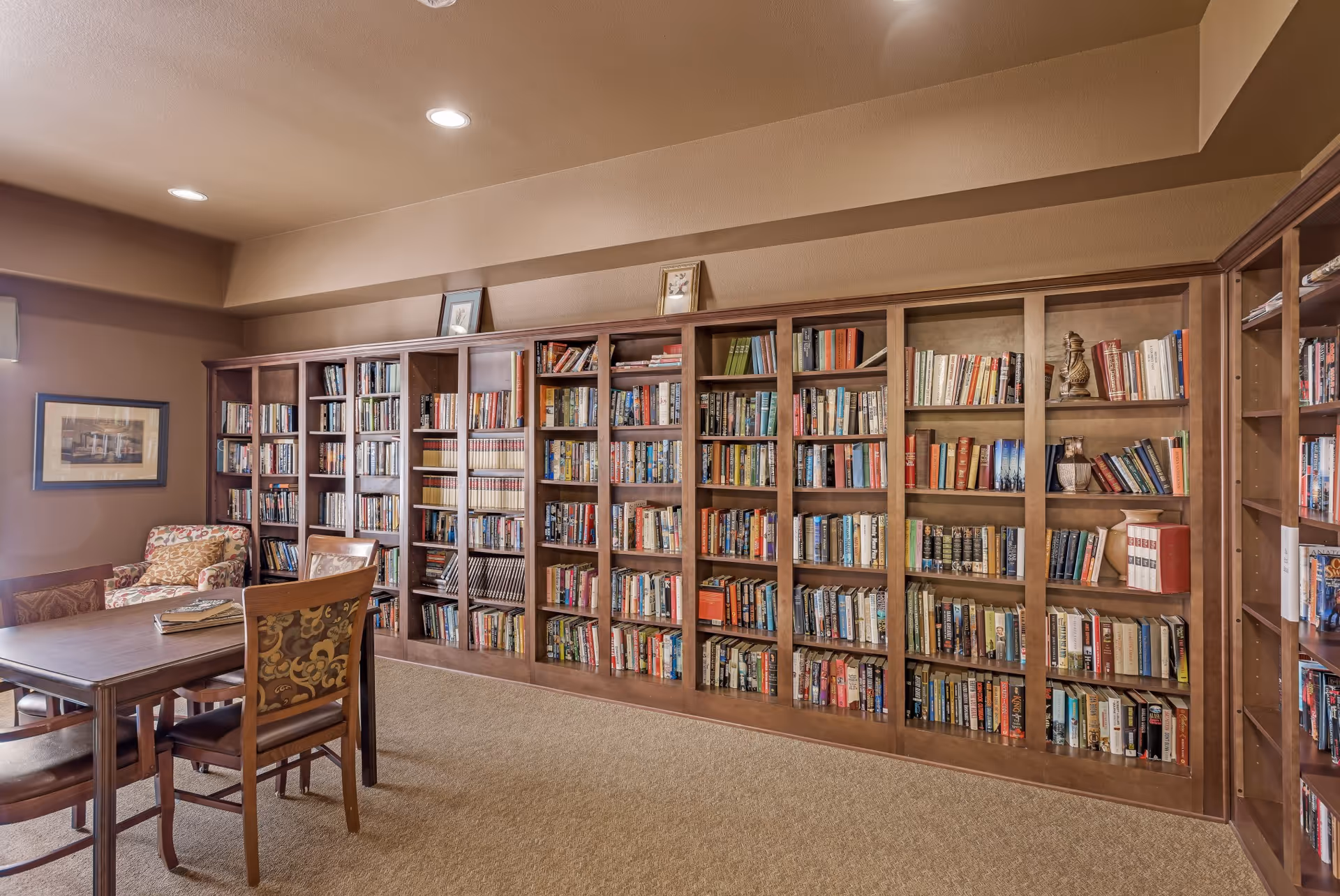 A cozy library room with wooden bookshelves filled with books along the wall. There is a wooden table with four chairs in the foreground and a patterned armchair in the corner. The room has warm beige walls and carpeted floor, with framed pictures on the wall and above the bookshelves.