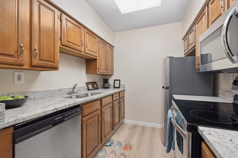 A narrow kitchen with wooden cabinets, granite countertops, a stainless steel dishwasher, refrigerator, microwave, and electric stove. There is a double sink, a coffee maker, and framed pictures on the countertop. The floor has light wood-style flooring and a colorful mat in front of the sink.
