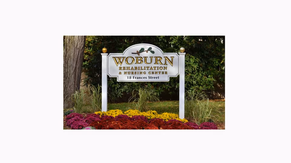 A white sign with gold and black lettering that reads 'Woburn Rehabilitation & Nursing Center, 18 Frances Street' surrounded by colorful flowers and greenery in an outdoor setting.