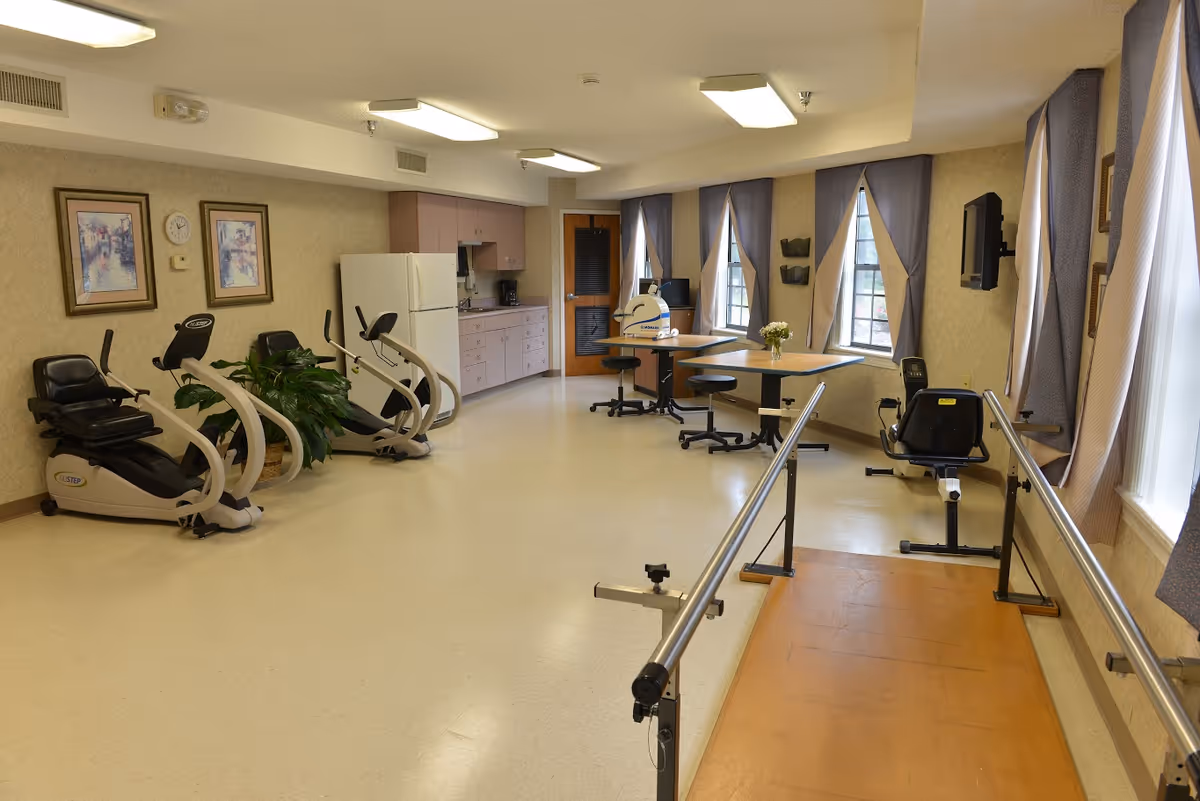 A spacious rehabilitation or exercise room with stationary exercise bikes, parallel bars for walking practice, tables with chairs, a refrigerator, cabinets, and windows with curtains letting in natural light.