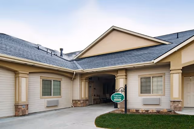 Front entrance of a single-story memory care building with a covered drive-through, columns, windows, and a small green sign on the lawn.