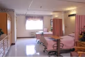 A hospital-style patient room with two beds separated by a pink privacy curtain. Each bed has a pink blanket and an adjustable overbed table. There is a window with floral curtains letting in natural light, a wooden dresser, and a wardrobe. The room has a clean, clinical appearance with light-colored walls and flooring.