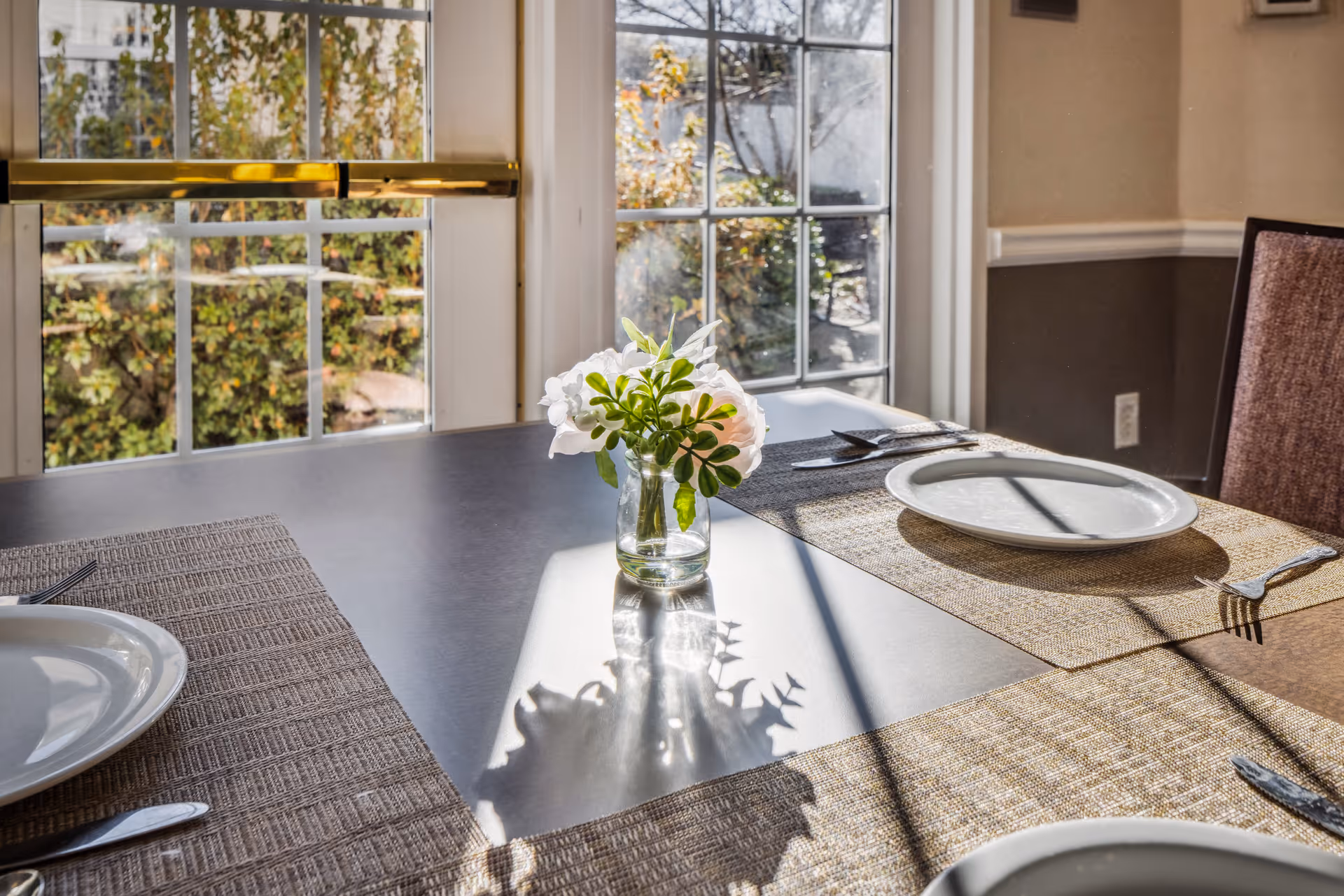 Sunlit dining table set with place settings and a small vase of flowers in front of a window.