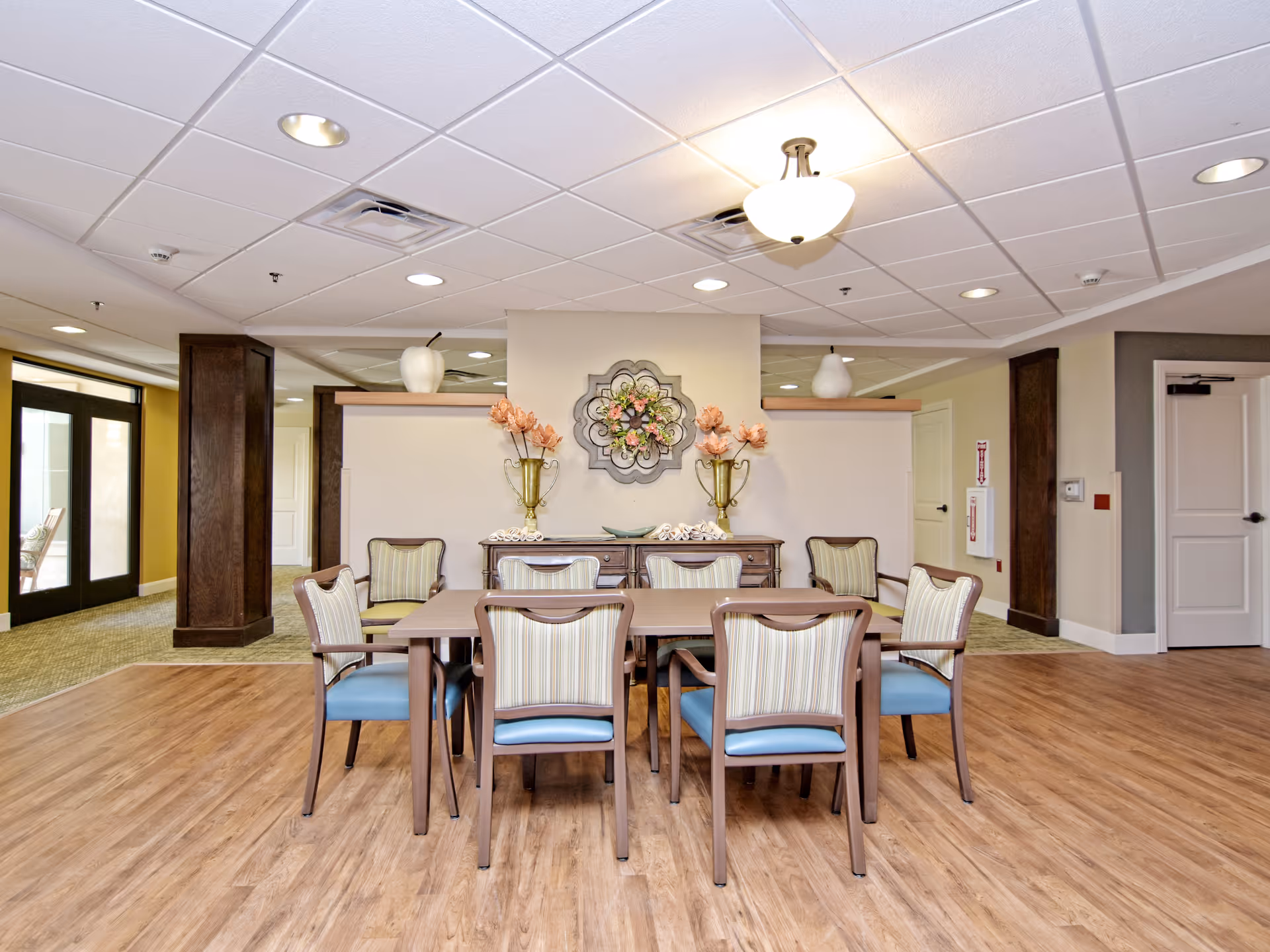 A dining area in a senior living facility with a rectangular wooden table surrounded by eight chairs with striped upholstery and blue seats. Behind the table is a wooden sideboard decorated with two gold vases holding pink flowers and a decorative wall piece with floral elements. The room has wood flooring, a drop ceiling with recessed lighting, and light-colored walls. There are doors and a hallway visible in the background.