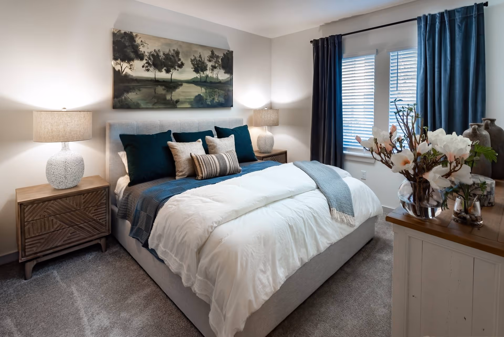 A cozy bedroom with a large bed featuring white and blue bedding and multiple pillows. There are two wooden nightstands with matching lamps on either side of the bed. A painting of trees over a body of water hangs above the bed. A window with blue curtains and white blinds is on the right side, and a wooden dresser with a glass vase of white flowers is in the foreground.