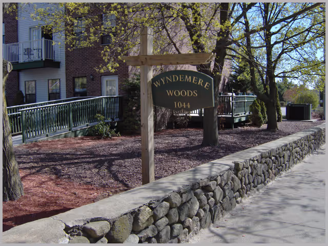 Outdoor view of the Wyndemere Woods facility sign mounted on a wooden post next to a stone wall. Behind the sign, there are trees with budding leaves and a brick building with balconies and a green railing ramp.