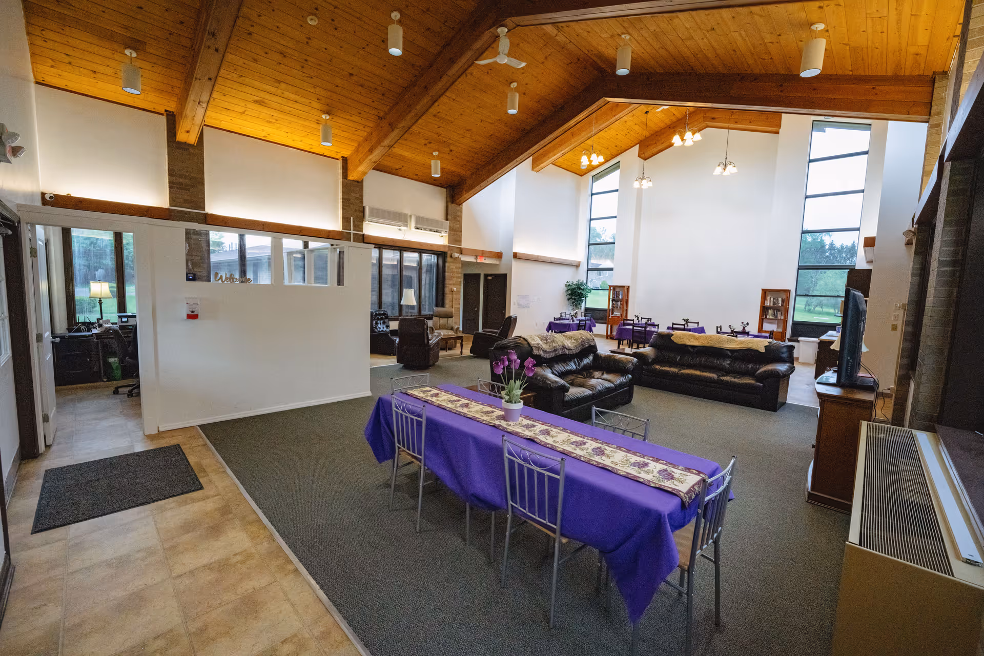 Spacious senior living common room with vaulted wooden ceiling, sofas and tables covered in purple tablecloths near large windows.