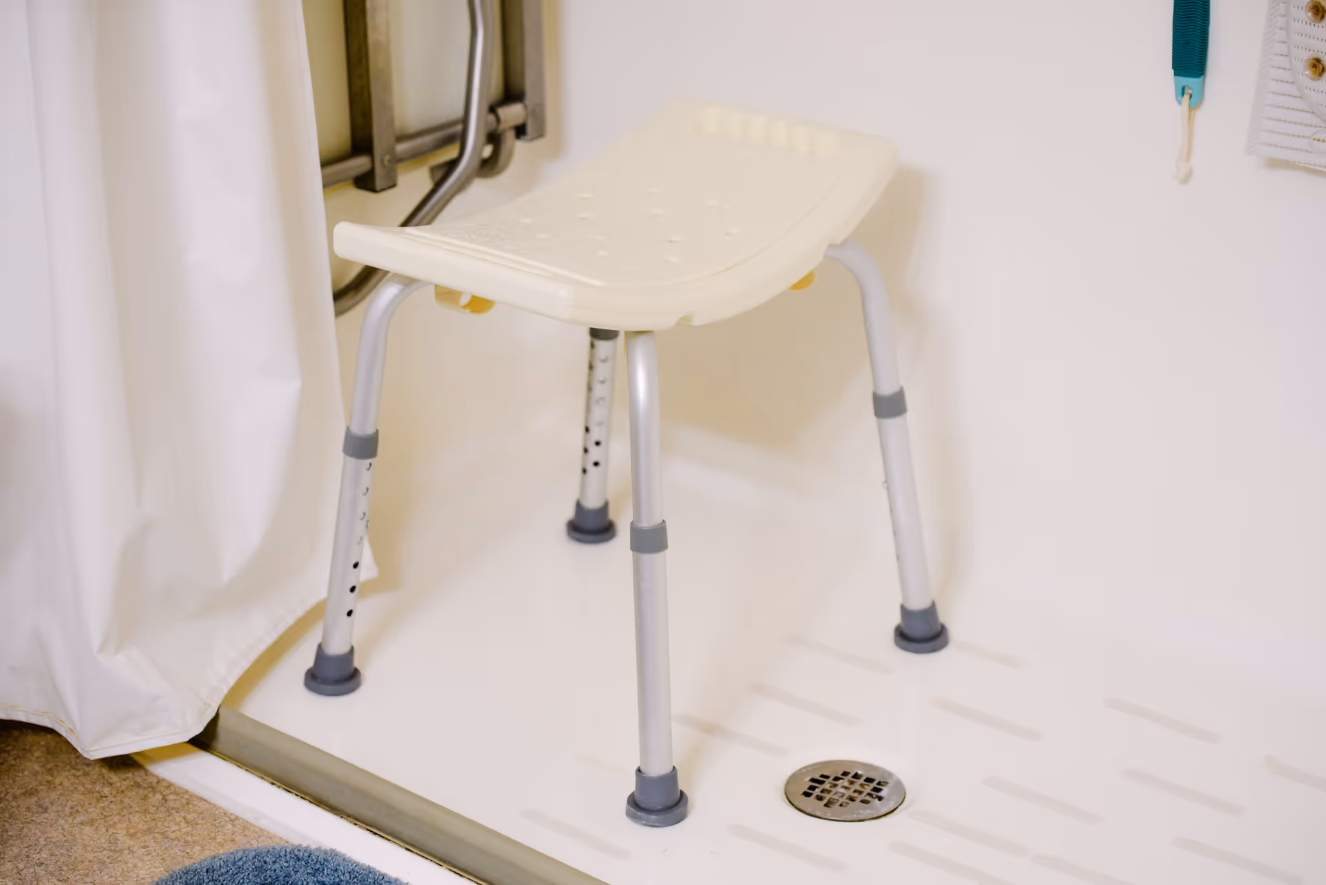 A white shower chair with metal legs inside a shower area, featuring a white shower curtain, a drain on the shower floor, and a blue bath mat outside the shower.