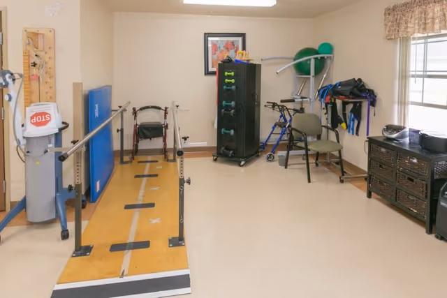 A rehabilitation therapy room with parallel bars for walking exercises, a walker, exercise equipment including dumbbells, exercise balls, and resistance bands, a chair, and a window with a curtain letting in natural light.