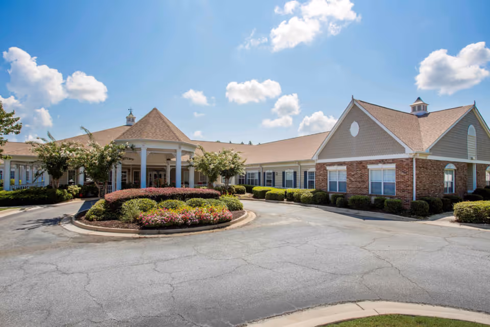 Exterior view of Oaks at Evans senior living facility showing a single-story building with a covered entrance, landscaped bushes and flowers, and a clear blue sky with scattered clouds.